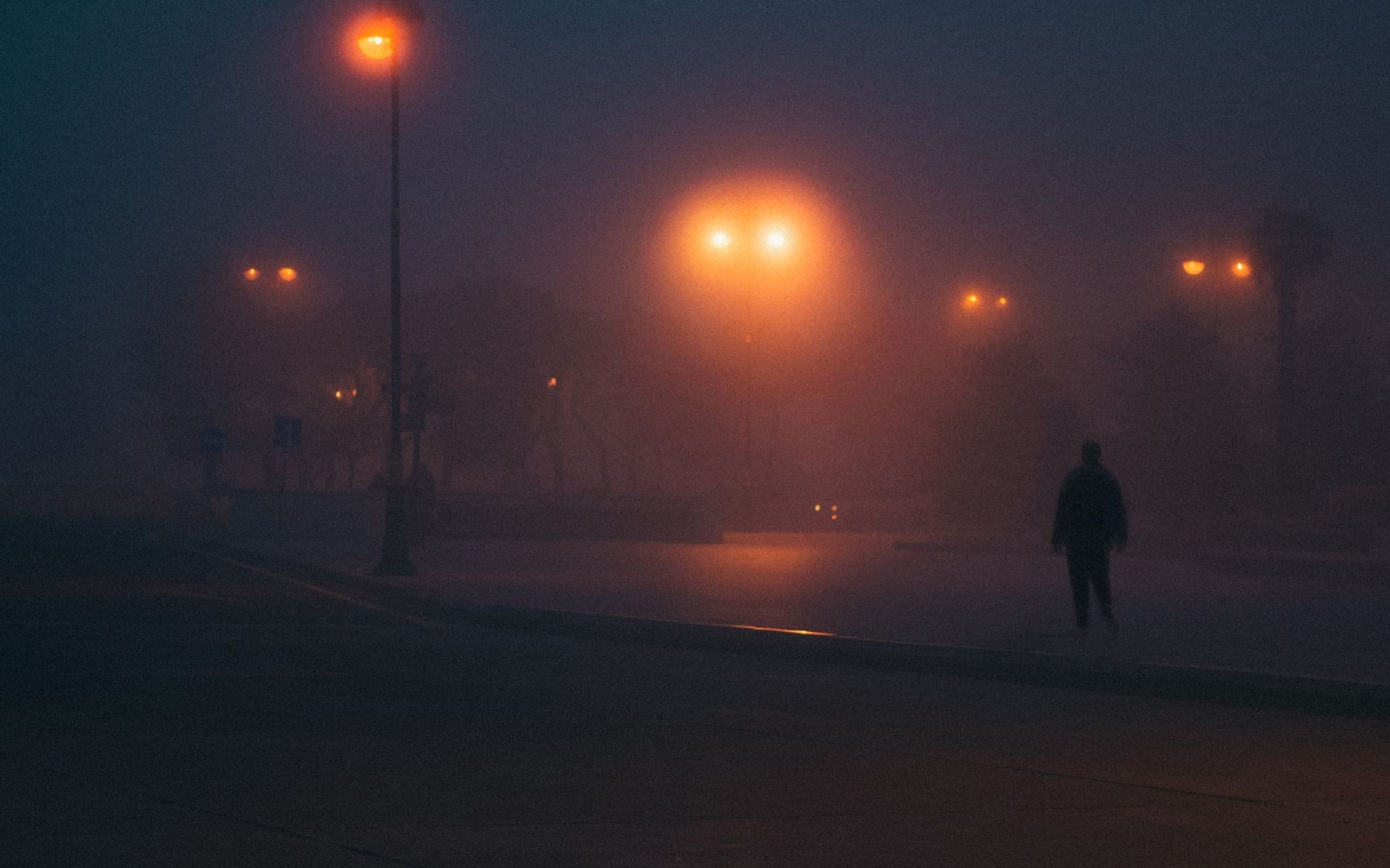 A lone figure walks along a foggy street lit by glowing streetlights at night