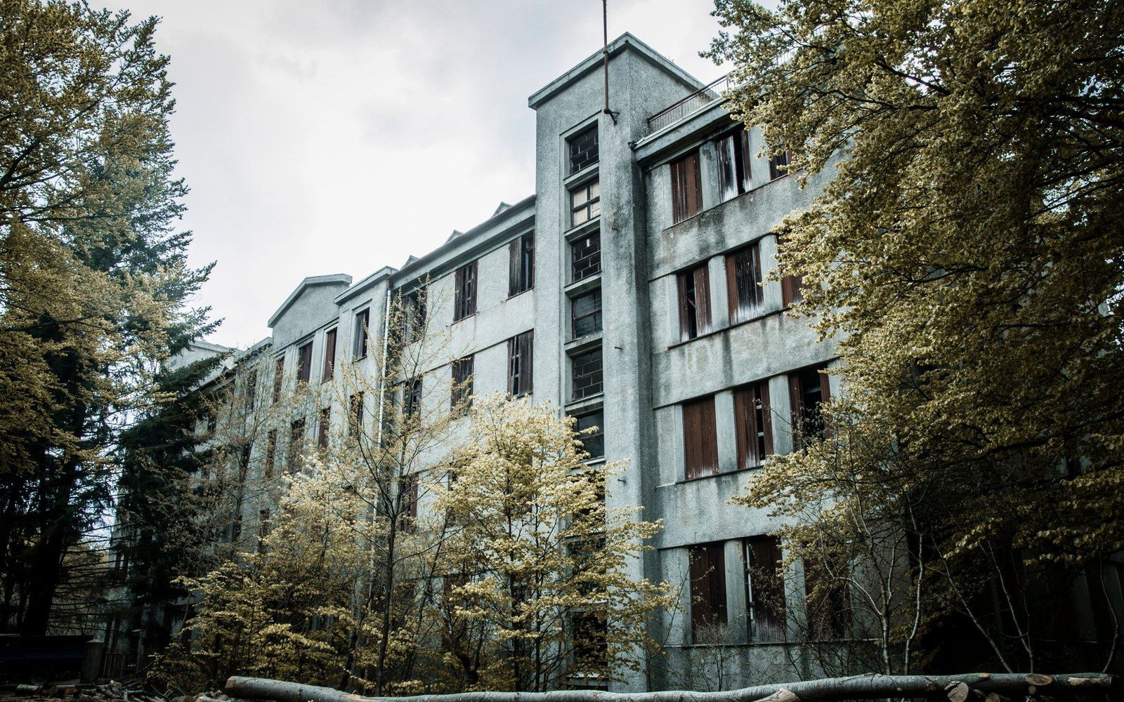 Abandoned multi-story sanatorium building with broken windows surrounded by overgrown trees