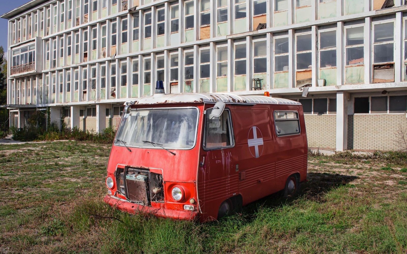 Rusting old ambulance with a red cross sits abandoned in front of a derelict multistory building